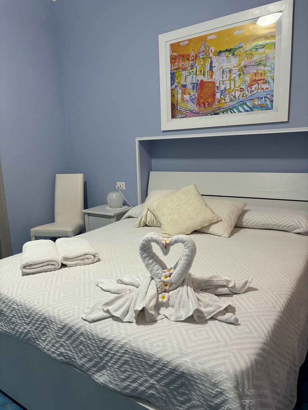 A neatly arranged bedroom featuring a white bed with decorative towel swans, colorful cityscape artwork on the wall, and modern furnishings in calming blue and white tones.