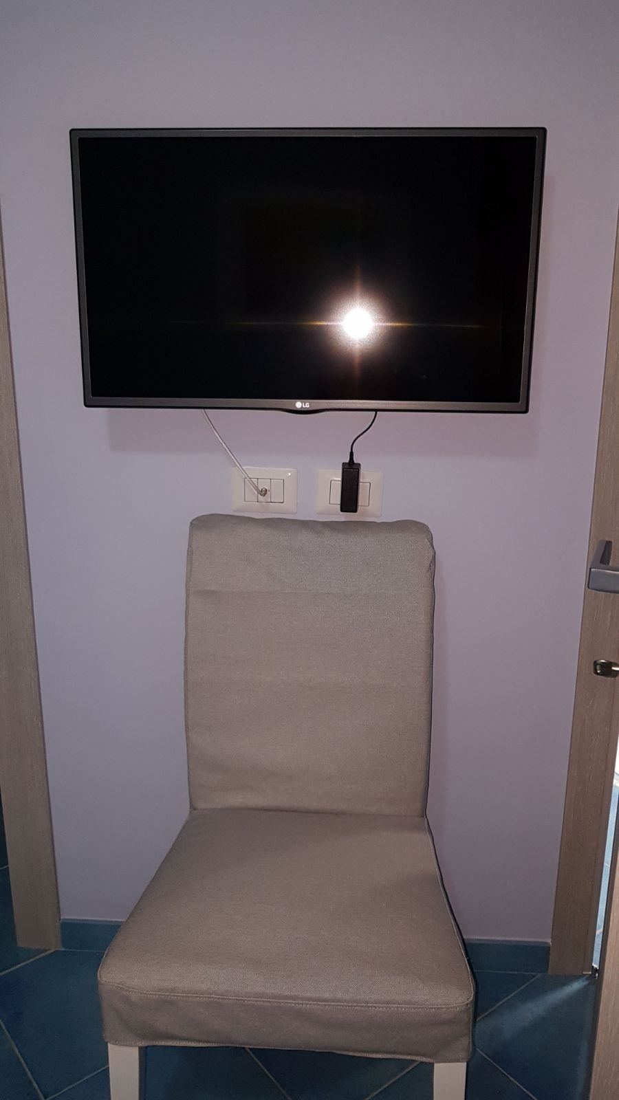 A small hotel room corner featuring a wall-mounted TV and a beige upholstered chair against a purple-painted wall.